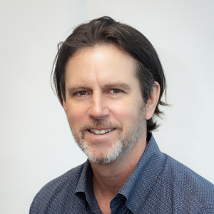 Portrait of a smiling man with brown hair and a light beard, wearing a blue patterned button-down shirt, looking at the camera.