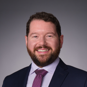 Portrait of a smiling man in a navy suit, lavender shirt, and purple patterned tie against a gray backdrop.